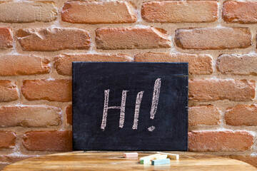 Black board with the word HI! drown by hand on wooden table on brick wall background.