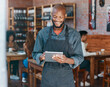 © Oostendorp/peopleimages.com - Young african american businessman wearing a apron working in a retail store using a digital tablet device. Portrait of a smiling small business owner, entrepreneur buying stock online using wireless