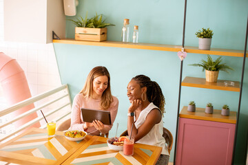 Wall Mural - Young black and caucasian woman having good time, drinking fresh juices and having healthy breakfast in the cafe