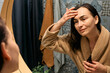 © Caterina Trimarchi - Middle-aged woman in bathrobe looking at mirror in bathroom and touching her face, applying hydrating anti-aging cosmetics on her skin and smiling.