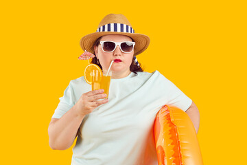 Fat woman enjoying cocktail at beach on summer holiday. Funny chubby lady in fedora sun hat and sunglasses isolated on yellow background holding inflatable ring and drinking cocktail. Vacation concept