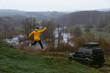 A man in a yellow jacket win jump. Off-road vehicle. A small compact car for extreme hobbies and recreation in wilderness. River and hills on the background. Mud dirt road. Gloomy rainy day.