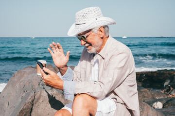 Wall Mural - Smiling Old Senior Man with Beard and White Hat Sitting at the Beach in Video Call by Smartphone - Vacation Retirement Lifestyle concept, Horizon over Water