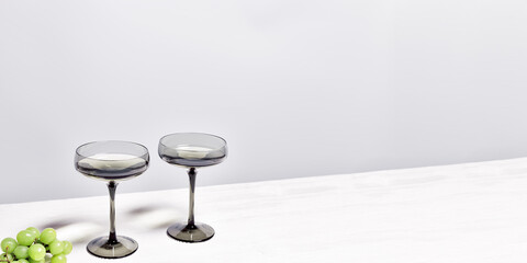 Gray shampagne glasses and grape om white wooden table, trend wine glass colored. Minimal summer still life with sparkling white wine in wide shampagne glass. Side view, copy space