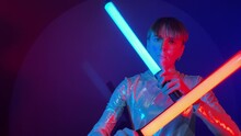 Concentrated Young Woman In Reflective Sparkling Costume Dancing With Neon Red Blue Tubes On Colorful Background. Female Person Makes Smooth Movements Illuminated Vibrant Colors. Futuristic Concept