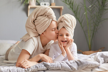 Motherhood, childhood, parenthood. Loving mother and daughter laying in bed wearing towel posing in bedroom at home, woman kissing her charming kid doing beauty procedures together.