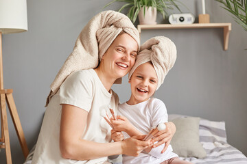 joyful woman hugging her little kid mother and daughter in towels posing in bedroom looking at camer