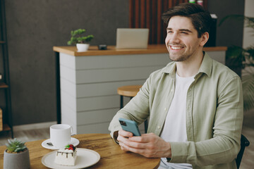Wall Mural - Young minded happy smiling caucasian man wears casual clothes sits alone at table in coffee shop cafe restaurant indoors work hold use mobile cell phone look aside rest relax during free time inside.