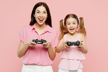 Wall Mural - Happy woman wear casual clothes with child kid girl 6-7 years old. Mother daughter hold in hand play pc game with joystick console isolated on plain pastel pink background. Family parent day concept.