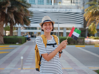 Wall Mural - Happy young asian female traveler with backpack and hat with UAE flag against scenic skyscrapers in Abu Dhabi. Immigration, student education and arab emirates citizenship concept