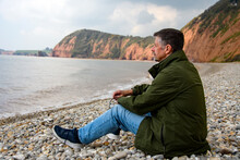 A Man Sits On A Beach In Front Of A Cliff, Staring Out To Sea. Concept Image, He Ponders His Retirement, Finances, Health And Pensions. He Is Isolated And Alone. Landscape View, Copy Space. Editorial.