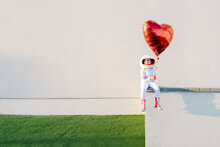 Girl Wearing Astronaut Costume Sitting On Wall Holding Heart Shape Balloon