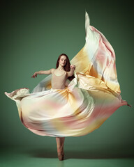 Wall Mural - Portrait with one young girl, beautiful ballerina wearing silk dress spinning in dance over dark green studio background