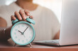© Kesinee - Asian woman's hand reaching for an alarm clock to check the time on desk with a computer laptop for the concept of work, study and time management.