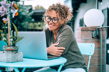 Happy woman using computer for video call outside home in the garden sitting on a table. Alternative office workplace outdoor. Modern people with laptop. Wireless online connection. People and leisure