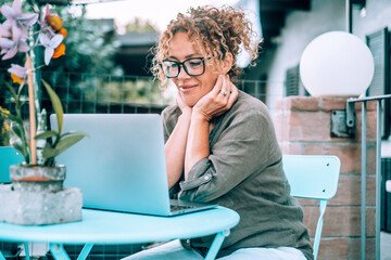 Wall Mural - Serene woman using computer outdoor home at the table enjoying outdoor technology online leisure activity. Modern lady watching laptop display and smile. People wearing glasses and relax lifestyle