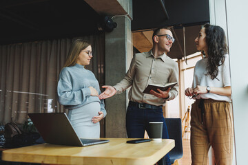 Modern pregnant woman working in office with colleagues. A young successful man helps a pregnant colleague at work. A beautiful pregnant woman works in the office. Modern pregnant woman