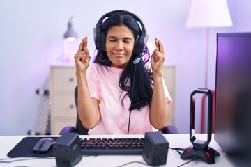 Poster - Mature hispanic woman playing video games at home gesturing finger crossed smiling with hope and eyes closed. luck and superstitious concept.