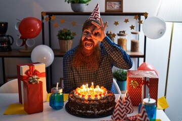 Canvas Print - Caucasian man with long beard celebrating birthday holding big chocolate cake smiling happy doing ok sign with hand on eye looking through fingers