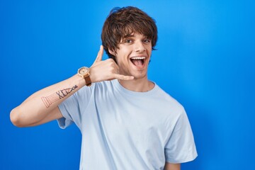 Poster - Hispanic young man standing over blue background smiling doing phone gesture with hand and fingers like talking on the telephone. communicating concepts.