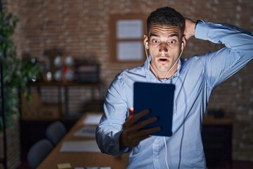 Wall Mural - Handsome hispanic man working at the office at night crazy and scared with hands on head, afraid and surprised of shock with open mouth