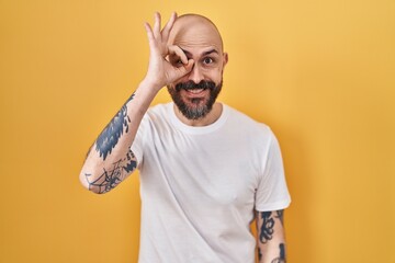 Wall Mural - Young hispanic man with tattoos standing over yellow background doing ok gesture with hand smiling, eye looking through fingers with happy face.