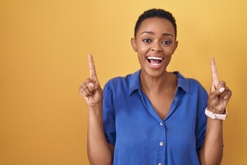 Sticker - African american woman standing over yellow background smiling amazed and surprised and pointing up with fingers and raised arms.