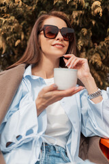 Sticker - Beautiful fashionable young girl in trendy stylish clothes with sunglasses sits in a cafe outdoors and drinks tea