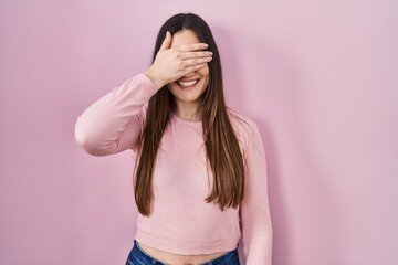 Canvas Print - Young brunette woman standing over pink background smiling and laughing with hand on face covering eyes for surprise. blind concept.