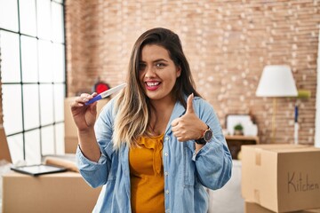 Wall Mural - Young hispanic woman holding pregnancy test result at new home smiling happy and positive, thumb up doing excellent and approval sign