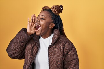 Sticker - African woman with braided hair standing over yellow background shouting and screaming loud to side with hand on mouth. communication concept.