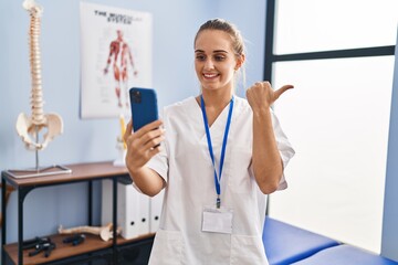 Poster - Young physiotherapist woman working at pain recovery clinic doing video appointment pointing thumb up to the side smiling happy with open mouth