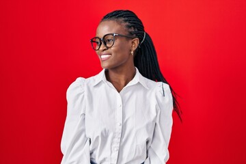 Sticker - African woman with braids standing over red background looking away to side with smile on face, natural expression. laughing confident.