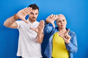 Canvas Print - Young brazilian mother and son standing over blue background doing frame using hands palms and fingers, camera perspective