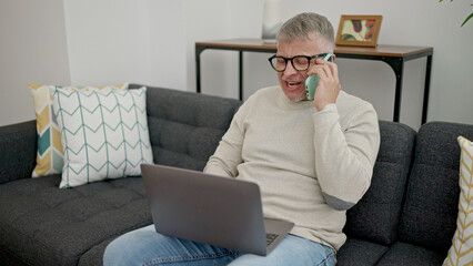 Sticker - Middle age grey-haired man talking on smartphone using laptop at home