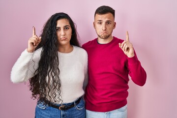 Wall Mural - Young hispanic couple standing over pink background pointing up looking sad and upset, indicating direction with fingers, unhappy and depressed.