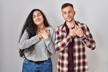 Poster - Young hispanic couple standing over white background smiling with hands on chest with closed eyes and grateful gesture on face. health concept.