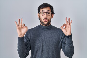 Canvas Print - Handsome latin man standing over isolated background looking surprised and shocked doing ok approval symbol with fingers. crazy expression