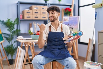 Poster - Arab man with beard painter sitting at art studio holding palette pointing thumb up to the side smiling happy with open mouth