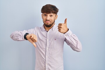 Sticker - Arab man with beard standing over blue background doing thumbs up and down, disagreement and agreement expression. crazy conflict