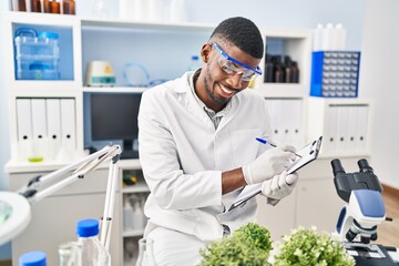 Sticker - Young african american man wearing scientist uniform writing on clipboard looking plant at laboratory