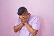 Young hispanic man standing over pink background with sad expression covering face with hands while crying. depression concept.
