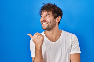 Poster - Hispanic young man standing over blue background smiling with happy face looking and pointing to the side with thumb up.