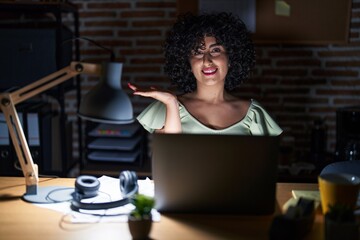 Poster - Young brunette woman with curly hair working at the office at night smiling cheerful presenting and pointing with palm of hand looking at the camera.