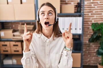 Sticker - Young caucasian woman working at small business ecommerce wearing headset amazed and surprised looking up and pointing with fingers and raised arms.