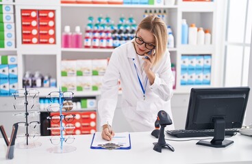 Canvas Print - Young blonde woman pharmacist write on document talking on smartphone at pharmacy