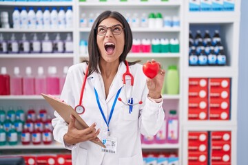 Poster - Brunette young woman working at pharmacy drugstore holding red heart angry and mad screaming frustrated and furious, shouting with anger looking up.