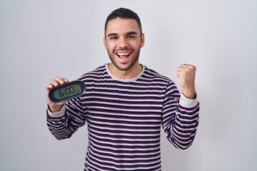 Poster - Young hispanic man wearing pyjama holding alarm clock screaming proud, celebrating victory and success very excited with raised arms