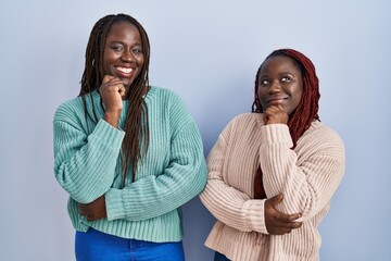 Poster - Two african woman standing over blue background with hand on chin thinking about question, pensive expression. smiling and thoughtful face. doubt concept.