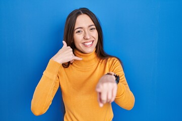 Sticker - Young brunette woman standing over blue background smiling doing talking on the telephone gesture and pointing to you. call me.